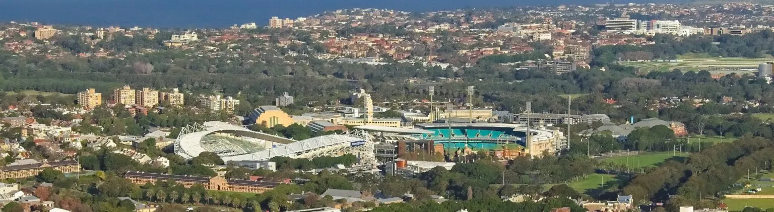 Aerial view of two large sports stadiums surrounded by parkland, with residential suburbs and the ocean beyond